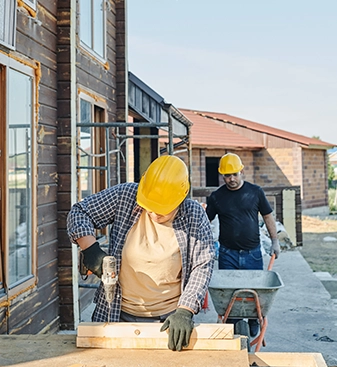 Mann im Vordergrund bohrt mit der Bohrmaschine ein Loch in eine Holzstaffel, Mann im Hintergrund schiebt eine Schreibtruhe vor sich her, sie arbeiten auf einer Baustelle und renovieren ein Holzhaus