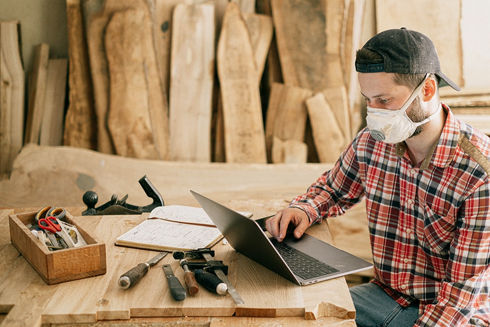 Handwerker mit Maske sitzt vor dem Laptop, vor ihm liegen auch Werkzeuge zur Bearbeitung von Holz auf der Werkbank. 