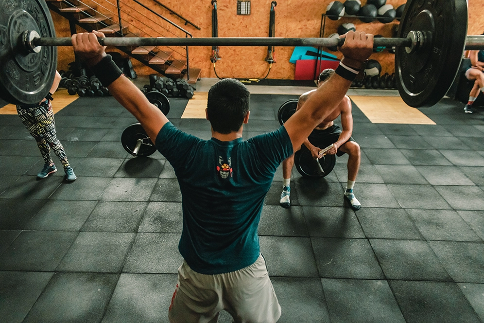 Mann in Rückenansicht stemmt Langhantel mit Gewichtsscheiben in die Höhe, Fitnesscoach sitzt vor ihm mit Notizbuch. 